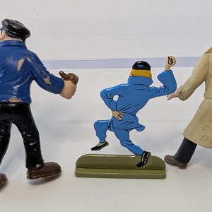 Puede incluir: Tres figuras vintage de Tint&iacute;n. Una figura lleva una chaqueta azul y pantalones negros, otra un mono azul, y la tercera un abrigo beige. Las figuras est&aacute;n sobre una superficie blanca.
