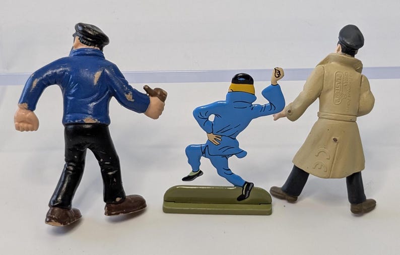 Puede incluir: Tres figuras vintage de Tint&iacute;n. Una figura lleva una chaqueta azul y pantalones negros, otra un mono azul, y la tercera un abrigo beige. Las figuras est&aacute;n sobre una superficie blanca.