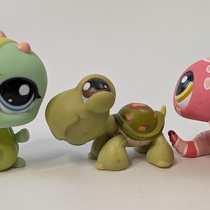 Puede incluir: Tres figuras coleccionables coloridas: una criatura verde parecida a un lagarto, una tortuga verde y una figura rosa con forma de hongo. Cada una tiene ojos grandes y expresivos y diseños únicos.