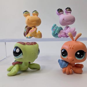 May include: Four colorful Littlest Pet Shop figurines. The toys include a light orange snail, a pink snail, a green turtle, and an orange ant. Each toy has large, expressive eyes and is made of plastic.