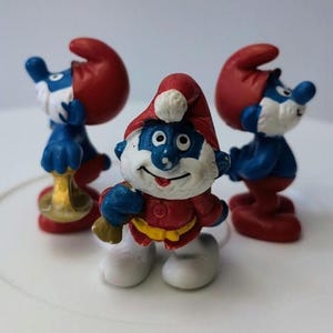 May include: Three Smurf figurines, one in a red and white Santa hat holding a gold trumpet, one in a red hat with a blue body, and one in a red hat with a blue body holding a gold trumpet.