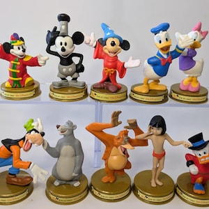 May include: A collection of Disney character figurines, including Mickey Mouse, Donald Duck, and Goofy. Each figurine is brightly colored and stands on a gold-colored base. The characters are in various poses, adding to the whimsical appeal of the collection.