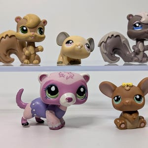 May include: Five small animal figurines in various colours. The collection includes a squirrel, a mouse, and two skunks. The figurines are made of plastic and have large, expressive eyes. The figurines are displayed on a clear shelf.