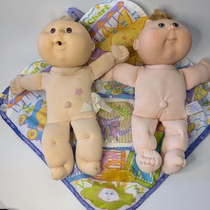 Peut inclure: Deux poupées Cabbage Patch Kids vintage avec des corps souples et des visages peints, posées sur un tapis de jeu coloré. Les poupées ont les bras et les jambes ouverts, l'une d'elles portant une étoile sur la poitrine. Une sucette est visible.