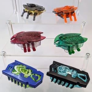 May include: Six colorful plastic toy bugs on a clear acrylic shelf. The bugs are translucent and have intricate details. The bugs are in various colors, including red, green, black, and blue.