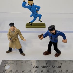 Puede incluir: Tres figuras en miniatura de personajes de c&oacute;mic. Una figura viste un traje azul, otra un abrigo color canela y la tercera una camisa azul y pantalones negros. Una regla de acero inoxidable est&aacute; en primer plano.