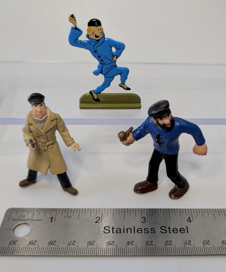 Puede incluir: Tres figuras en miniatura de personajes de c&oacute;mic. Una figura viste un traje azul, otra un abrigo color canela y la tercera una camisa azul y pantalones negros. Una regla de acero inoxidable est&aacute; en primer plano.