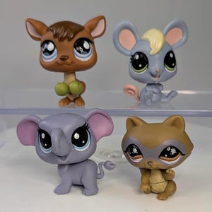 Könnte beinhalten: Vier farbenfrohe, sammelbare Tierfiguren. Das Set enthält ein braunes Reh mit grünen Akzenten, eine graue Maus mit einem gelben Büschel, einen lila Elefanten mit rosa Ohren und einen braunen Waschbären mit grauer Maske.