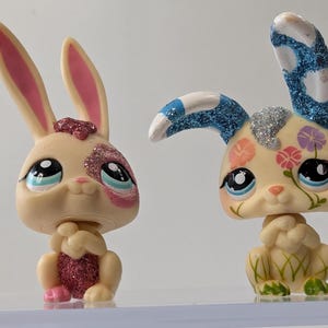 Puede incluir: Dos figuras decorativas de conejos. Uno tiene orejas rosas y un parche rosa brillante sobre el ojo. El otro tiene purpurina azul en las orejas y diseños florales en la cabeza. Ambos tienen ojos grandes.