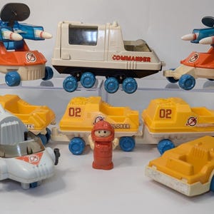 Peut inclure: Ensemble de véhicules jouets vintage de différentes conceptions et couleurs. Comprend un rover spatial blanc et orange, un camion blanc avec le texte "COMMANDER", des remorques jaunes avec le numéro "02" et une figurine d'astronaute rouge. Les jouets sont sur une étagère en plastique transparent.