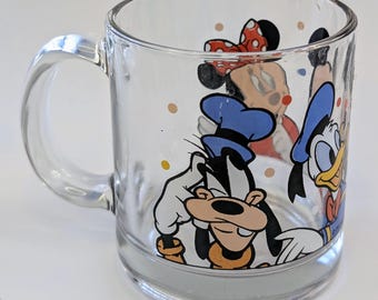 May include: A clear glass mug with a handle features a colorful cartoon design of Mickey Mouse, Minnie Mouse, Donald Duck, and Goofy. The characters are surrounded by yellow and orange polka dots.