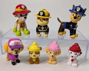 May include: A collection of seven Paw Patrol character figurines. The figurines are of various dog breeds, each wearing a uniform and hat in different colors, including red, yellow, blue, and pink. The figurines are displayed on a clear, elevated platform.