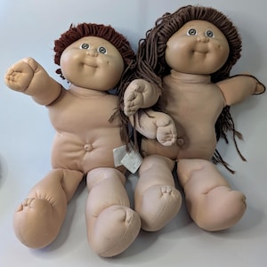 Può includere: Due bambole vintage Cabbage Patch Kids. Le bambole hanno corpi morbidi color carne, con mani e piedi dettagliati. Una bambola ha capelli di filo marrone, e l'altra ha capelli di filo marrone scuro. Entrambe le bambole hanno facce sorridenti e sono sedute.