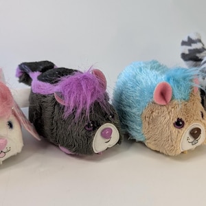 Peut inclure: Quatre animaux en peluche alignés. Le premier est blanc avec des détails roses, le second est noir et violet, le troisième est beige et bleu, et le quatrième est gris et blanc rayé avec des détails roses. Chaque animal a un design unique.