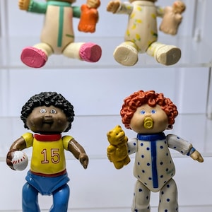 Puede incluir: Cuatro muñecas Cabbage Patch Kids, dos con pijamas, una con uniforme de béisbol con el número 15 y otra con un pijama azul y blanco de lunares.