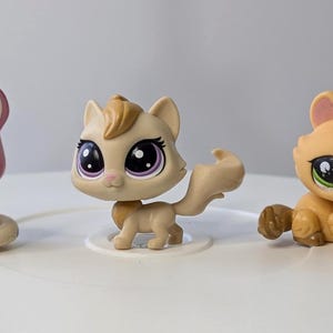Op de afbeelding: Drie kleine plastic kattenfiguren. De kat links is roze en wit met blauwe ogen. De kat in het midden is beige met bruin en wit haar en paarse ogen. De kat rechts is oranje met groene ogen.