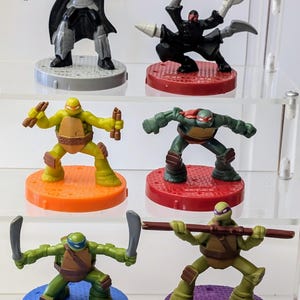 Könnte beinhalten: Sechs Teenage Mutant Ninja Turtles Actionfiguren, darunter Leonardo, Donatello, Raphael, Michelangelo, Shredder und ein Fußsoldat. Jede Figur ist in Kampfstellung und steht auf einem bunten Sockel.