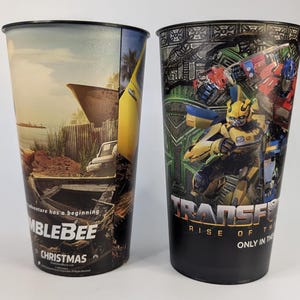 May include: Two tall plastic cups. One cup features a scene with a boat, a car, and the text "BUMBLEBEE CHRISTMAS". The other cup has a Transformers graphic with the text "TRANSFORMERS RISE OF THE BEASTS ONLY IN THEATERS".