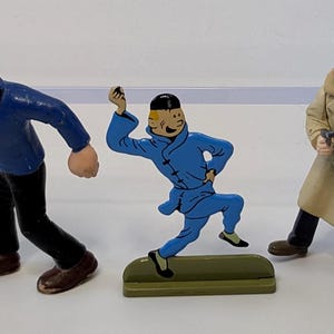 Puede incluir: Tres figuras de personajes de una serie de c&oacute;mics. Las figuras incluyen un marinero con una camisa azul, un personaje con un mono azul y un hombre con un abrigo color beige. Las figuras est&aacute;n sobre una superficie blanca.