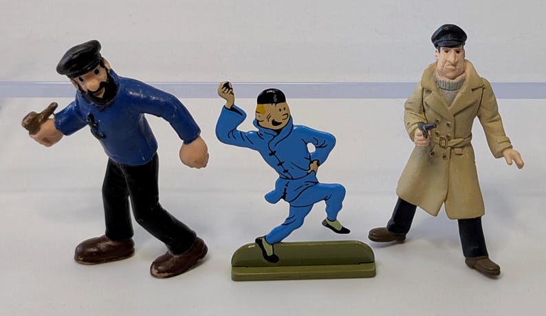 Puede incluir: Tres figuras de personajes de una serie de c&oacute;mics. Las figuras incluyen un marinero con una camisa azul, un personaje con un mono azul y un hombre con un abrigo color beige. Las figuras est&aacute;n sobre una superficie blanca.