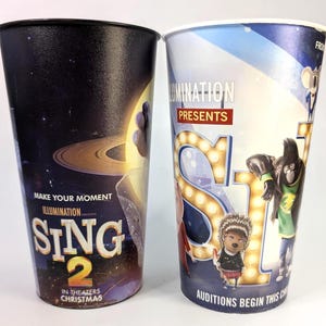 Op de afbeelding: Twee grote, kleurrijke papieren bekers met promotionele afbeeldingen voor de animatiefilm "Sing 2". Eén beker heeft een donkere achtergrond met de titel van de film en de tekst "Make Your Moment". De andere beker heeft een lichtblauwe achtergrond met de titel en de personages van de film.