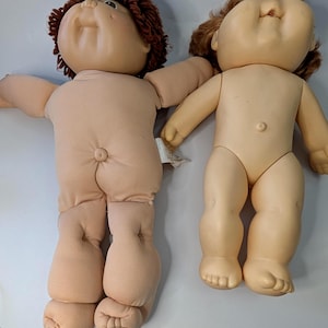 Peut inclure: Deux poupées Cabbage Patch Kids vintage. L'une a un corps en tissu souple et l'autre un corps en plastique. Les deux poupées ont des cheveux bouclés bruns et un teint neutre.