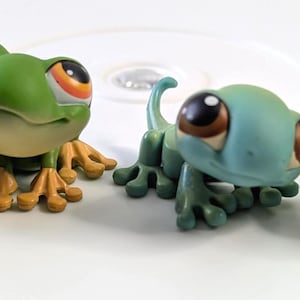 May include: Four toy reptile figurines in various colours. The toys include a green frog with orange and yellow accents, a light blue lizard, and two green turtle-like figures. The toys have large eyes and are posed on a white surface.