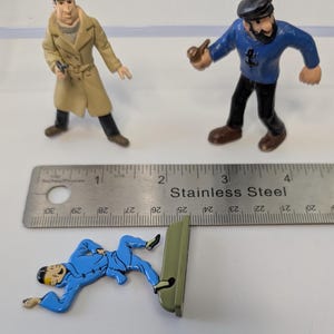 Puede incluir: Tres figuras de Tint&iacute;n: un detective con abrigo beige, un capit&aacute;n con pipa y Tint&iacute;n con un traje azul. Una regla de acero inoxidable est&aacute; en la imagen. Las figuras miden aproximadamente 2,5 a 5 cm de altura.