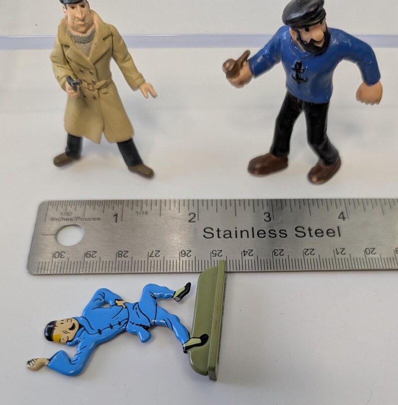 Puede incluir: Tres figuras de Tint&iacute;n: un detective con abrigo beige, un capit&aacute;n con pipa y Tint&iacute;n con un traje azul. Una regla de acero inoxidable est&aacute; en la imagen. Las figuras miden aproximadamente 2,5 a 5 cm de altura.