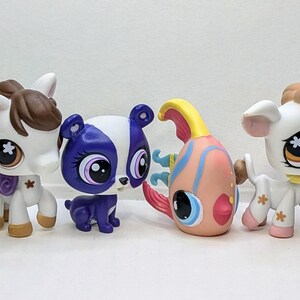 May include: A collection of seven small plastic toy animals. The animals include a grey skunk, a white horse with brown hair, a purple and white dog, a pink and orange fish, a white cow with brown hair, a brown squirrel, and a brown and white cat.