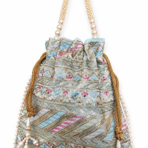 May include: A light blue drawstring bag with a floral and geometric pattern in pink, gold, and blue. The bag has a beaded handle and a beaded trim along the bottom. The drawstring is gold with a pearl tassel.