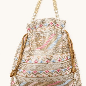 May include: An intricately embroidered drawstring bag with a cream-colored base, adorned with pink, blue, and gold threadwork. The bag features pearl-beaded handles and a pearl-trimmed drawstring closure. The bag's design includes floral and geometric patterns.