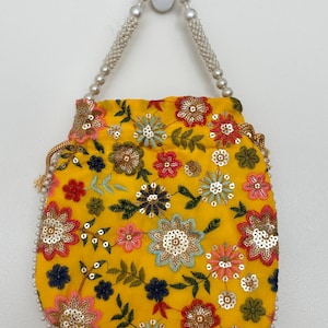 May include: Yellow embroidered bag with floral patterns in red, blue, pink, and gold sequins. The bag has a drawstring closure and pearl beaded handles. The bottom edge is also trimmed with pearls. The bag is hanging against a white wall.