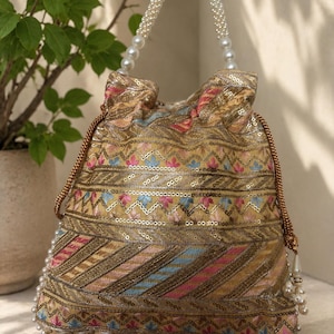 May include: An ornate, gold-toned drawstring bag with intricate embroidery in pink, blue, and gold. The bag features a beaded handle and a beaded base, creating a luxurious look. The design includes sequins and a tassel detail.