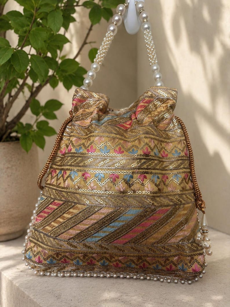 May include: An ornate, gold-toned drawstring bag with intricate embroidery in pink, blue, and gold. The bag features a beaded handle and a beaded base, creating a luxurious look. The design includes sequins and a tassel detail.