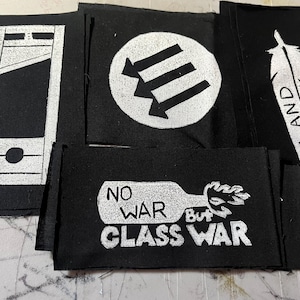 May include: Four black fabric patches with white designs. The patches feature a guillotine, three arrows, a feather with the words "Land Back", and a Molotov cocktail with the words "No War But Class War".