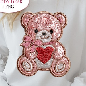 Op de afbeelding: Witte sweatshirt met een roze pailletten teddybeer ontwerp met een rood hart. De teddybeer heeft een roze strik en is omlijnd in een donkerdere roze kleur. De tekst "Teddy Bear 1 PNG" staat in een witte ovaal.