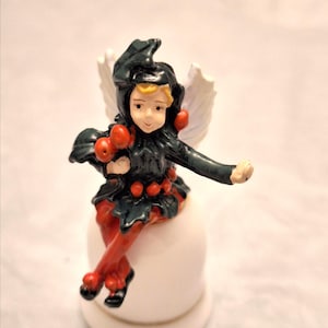 May include: A small figurine of a winged character wearing a dark green hooded outfit with red berry accents, sitting atop a smooth white rounded base. The character has blonde hair, a light complexion, and white wings extending from the back.