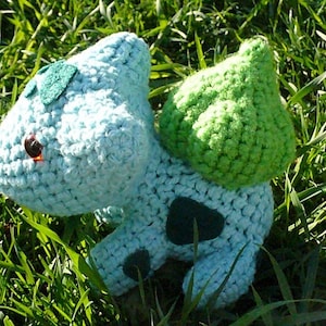 May include: A handmade crochet plush toy in the shape of a Bulbasaur. The toy is primarily light blue with a green bulb on its back. It has dark green spots and leaves, and a brown button eye. The plush is set in a bed of green grass.