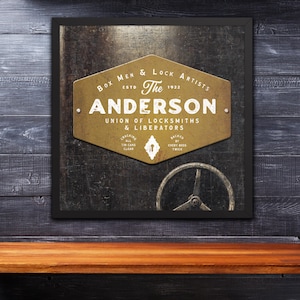 Puede incluir: Cartel enmarcado de estilo vintage con el texto "The Anderson Union of Locksmiths & Liberators" en blanco y dorado. El cartel incluye un gráfico de volante y está colocado sobre un fondo texturizado.