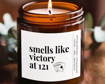 May include: A lit candle in a brown glass jar with a white label that reads "smells like victory at 121" and a playing card graphic. The candle sits on a wooden table with greenery.