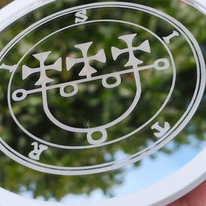 Peut inclure: Un miroir circulaire avec un cadre blanc présentant un motif de sceau gravé en blanc au centre. Le sceau se compose de trois symboles de croix disposés horizontalement au-dessus d'une ligne courbe, entourés d'une bordure circulaire avec les lettres S, I, R, T.