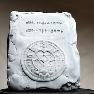 May include: A rectangular white stone-like tablet with an uneven, textured border. The center features a circular relief carving of a geometric star, wings, and celestial symbols. Above the circle, two lines of small, etched, unidentifiable characters.