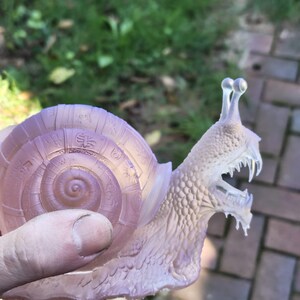 Può includere: Una statuetta di lumaca rosa pallido traslucida caratterizzata da un guscio a spirale dettagliato con vari simboli circolari e un corpo strutturato con la bocca aperta che rivela denti affilati e due peduncoli oculari.