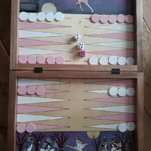 May include: A wooden backgammon game board with a painted scene of dancers and musicians. The board is open and shows the playing surface with pink and white checkers. Two dice are on the board, one showing a 3 and the other showing a 5.
