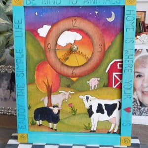 May include: A wooden clock with a painted scene of a farm with a red barn, a cow, a pig, a goat, and a black sheep. The clock face is a circle with a sun and clouds painted on it. The clock is surrounded by a turquoise frame with the words "Be Kind to Animals" and "Home is Where Your Heart Is" painted on it. The clock is sitting on a black and white checkered base.