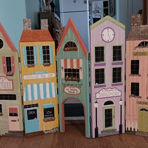 May include: A decorative wooden room divider featuring five colorful, hand-painted buildings. Each building has unique details like windows, doors, and signs, including "Gracie's Boutique," "Catch of the Day," and "Wisteria Inn." The divider is designed to resemble a charming village scene.