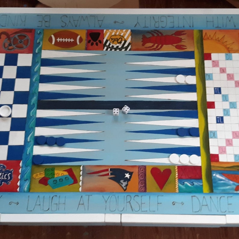 K&ouml;nnte beinhalten: Ein blaues und wei&szlig;es Backgammon-Spielbrett aus Holz mit einem Schachbrett auf der einen Seite und einem Backgammon-Brett auf der anderen. Das Brett hat eine Vielzahl von bunten Illustrationen, darunter ein Fu&szlig;ball, ein Pfotenabdruck, ein Hummer, ein Herz und eine Welle. Das Brett hat auch den Text "With Integrity", "Always Be Kind", "Laugh At Yourself", und "Dance".