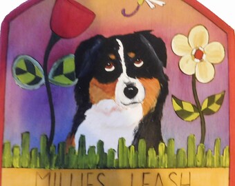 May include: Painted wooden sign featuring a dog with black, brown, and white fur, surrounded by flowers and grass. A red tulip and a yellow flower are on either side of the dog. The sign reads "MILLIES LEASH". A dragonfly is in the upper right corner.