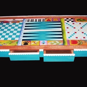 K&ouml;nnte beinhalten: Ein h&ouml;lzerner Spieltisch mit einer t&uuml;rkisblauen Tischplatte und braunen Beinen. Die Tischplatte zeigt ein Schachbrett, ein Backgammon-Brett und ein Tic-Tac-Toe-Brett. Der Tisch hat zwei Schubladen mit wei&szlig;en, gewellten Kanten. Der Tisch ist mit bunten Designs und den Worten "By With Hugs and Kisses Be Thankful Try New Things" bemalt.